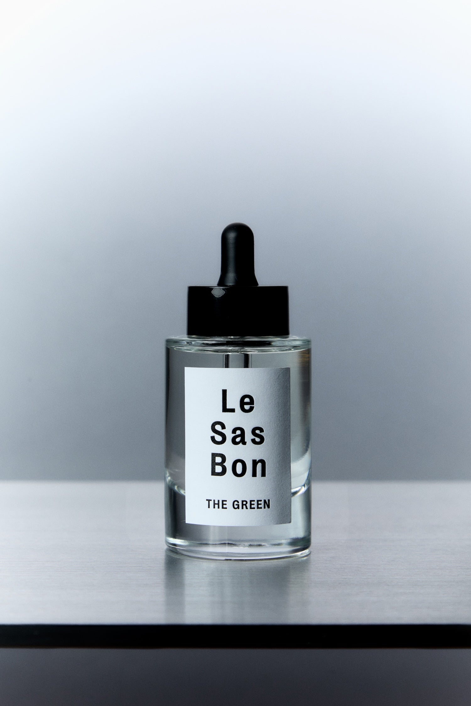 Clear cylinder dropper bottle with clear liquid of Le Sas Bon's "The Droppa" cruelty-free toilet fragrance. Bottle has a branded Le Sas Bon white sticker label. Product against grey gradient background