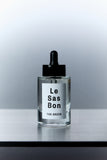Clear cylinder dropper bottle with clear liquid of Le Sas Bon's "The Droppa" cruelty-free toilet fragrance. Bottle has a branded Le Sas Bon white sticker label. Product against grey gradient background