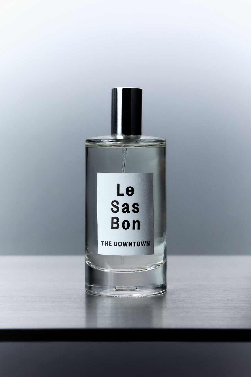 Cylinder glass spray bottle with Le Sas Bon "The Downtown" clear liquid room spray and branded label against grey gradient background 
