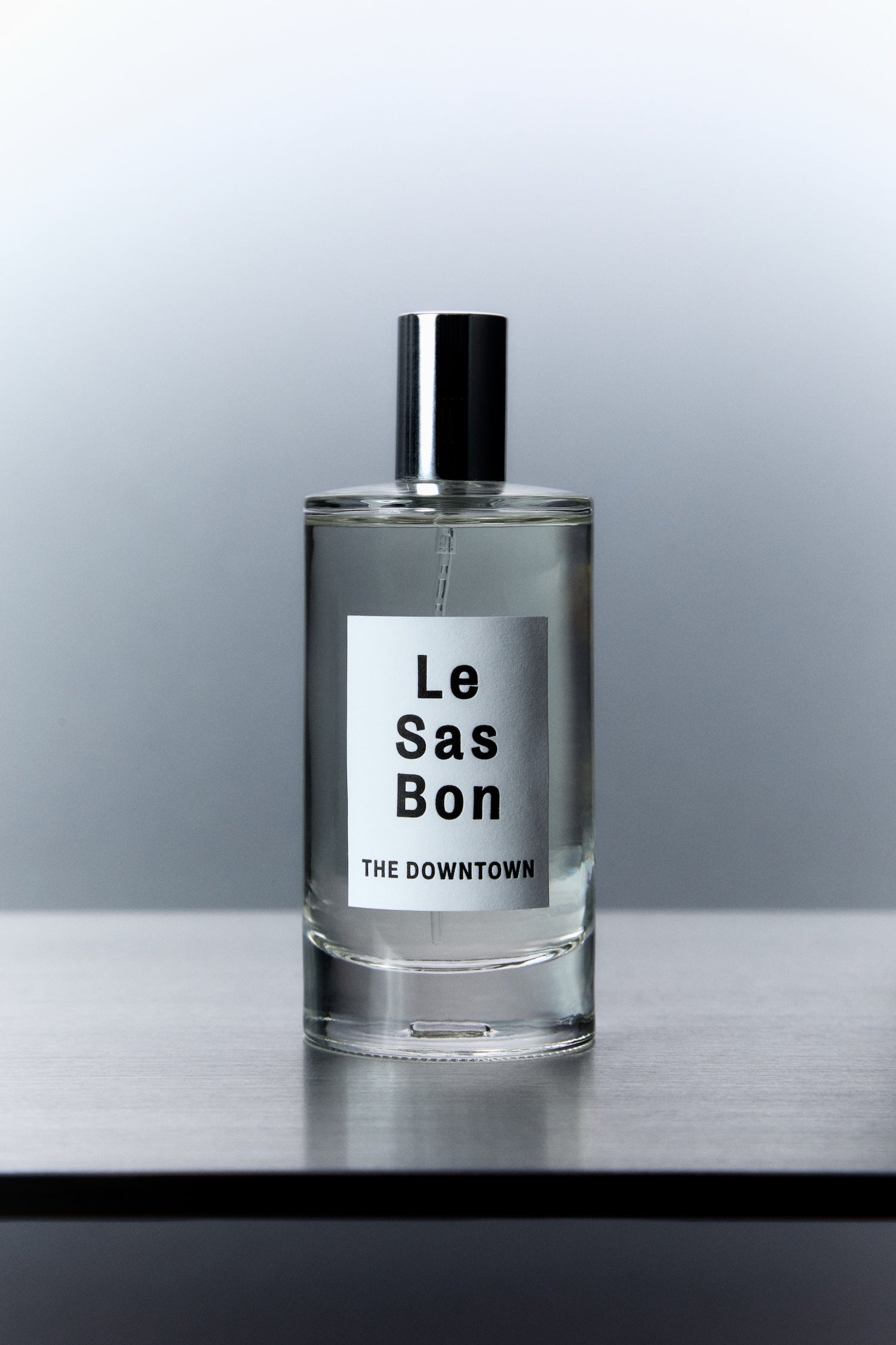 Cylinder glass spray bottle with Le Sas Bon "The Downtown" clear liquid room spray and branded label against grey gradient background 