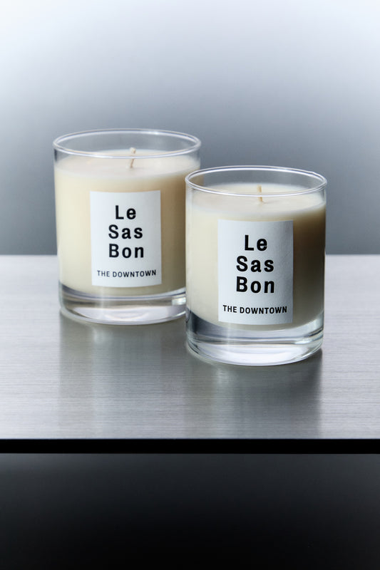 Le Sas Bon "The Downtown" natural and sustainable candle in 7.5oz and 10oz next to each other in glass vessels with white rectangle branded sticker against a silver background