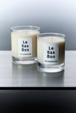 Le Sas Bon "The Downtown" natural and sustainable candle in 7.5oz and 10oz next to each other in glass vessels with white rectangle branded sticker against a silver background