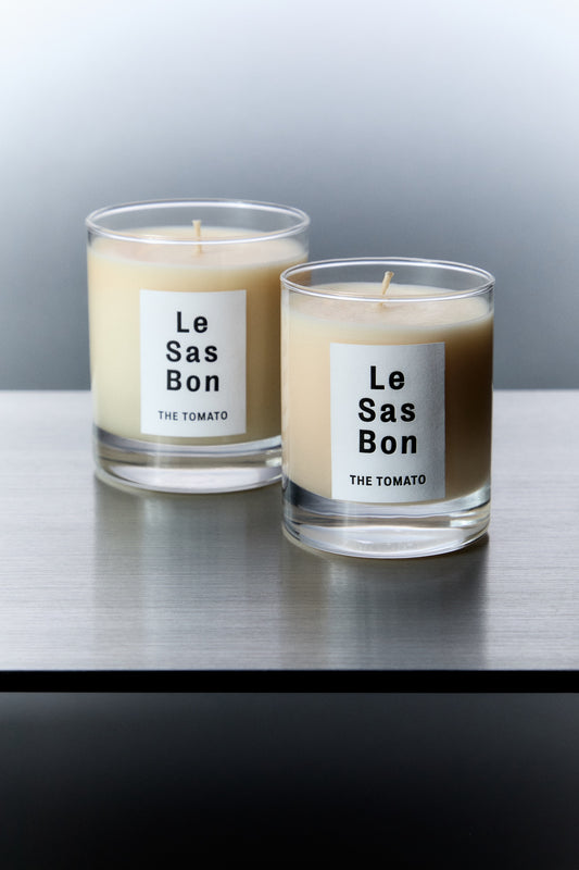 Le Sas Bon "The Tomato" coconut candle in 7.5oz and 10oz next to each other in glass vessels with white rectangle branded labels against a silver background